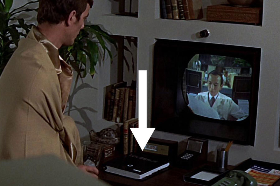 <p><strong>Figure 6.6</strong> In this scene in Forbin’s living quarters, we glimpse the logotype on the cover of hardbound documentation for Colossus, sitting on the desk below the video monitor.</p>