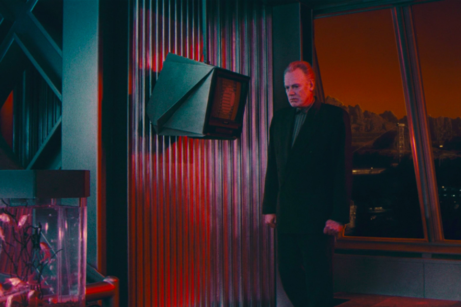 <p>Administrator Vilos Cohaagen brooding in front of a wall-mounted monitor displaying a one-color version of the Federal Colonies logo, with Mars’ red exterior glow cast over the scene.</p>