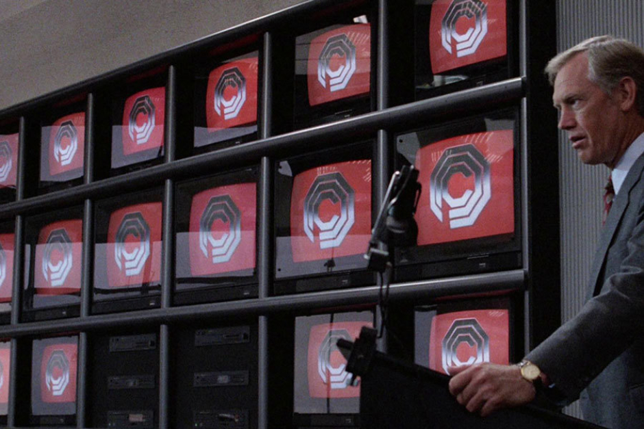 <p><strong>Figure 3.3</strong> As OCP Senior President Dick Jones addresses the boardroom, we see a screen version of the logo arrayed in metallic chrome on red.</p>