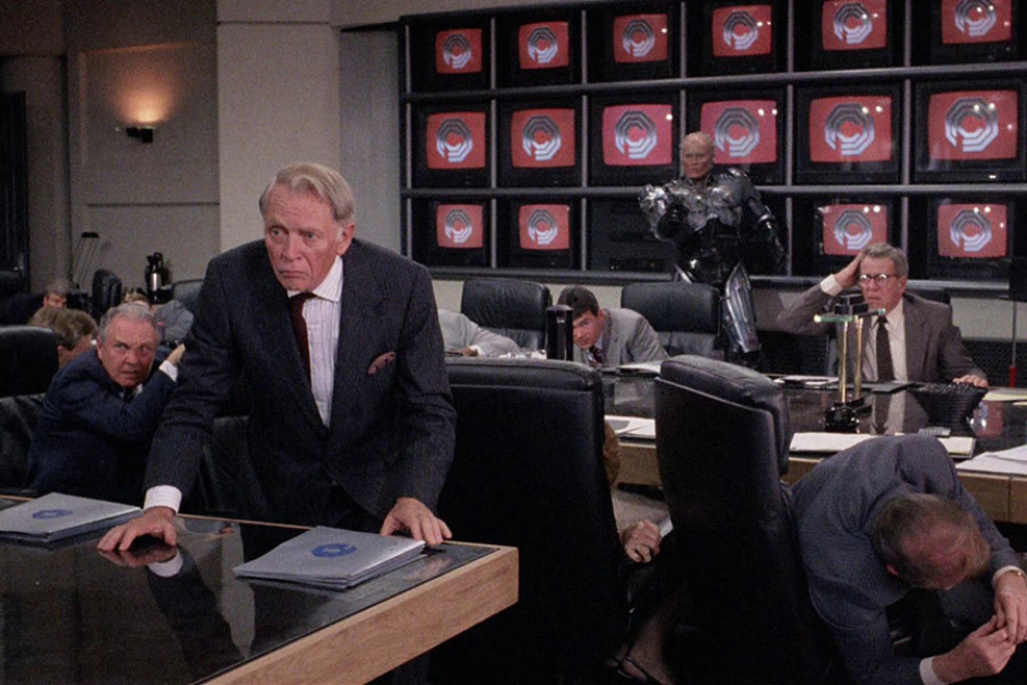 <p><strong>Figure 3.17</strong> The boardroom at OCP features a wall of television screens, which regularly display a beveled chrome OCP logo over a red background.</p>